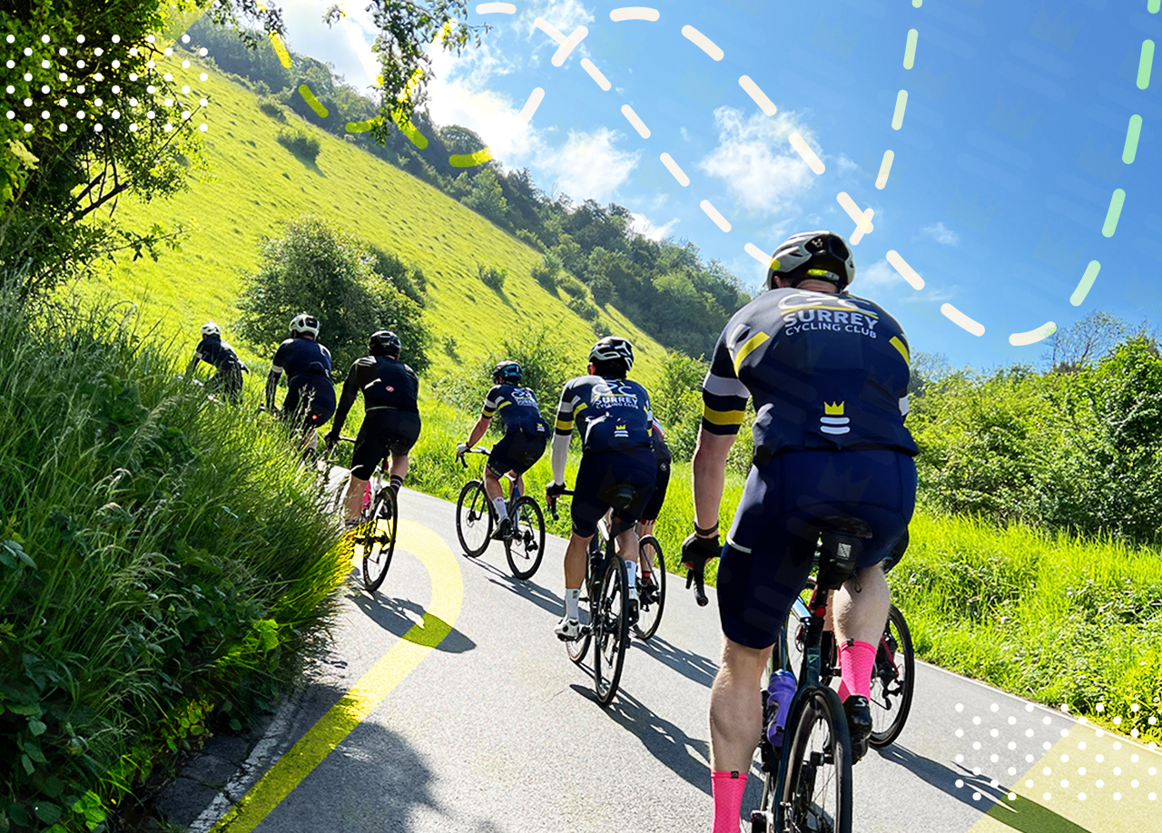 A group of cyclists from "Surrey Cycling Club" ride on a scenic, winding road with greenery on both sides. The cyclists are wearing matching navy-blue jerseys and helmets.