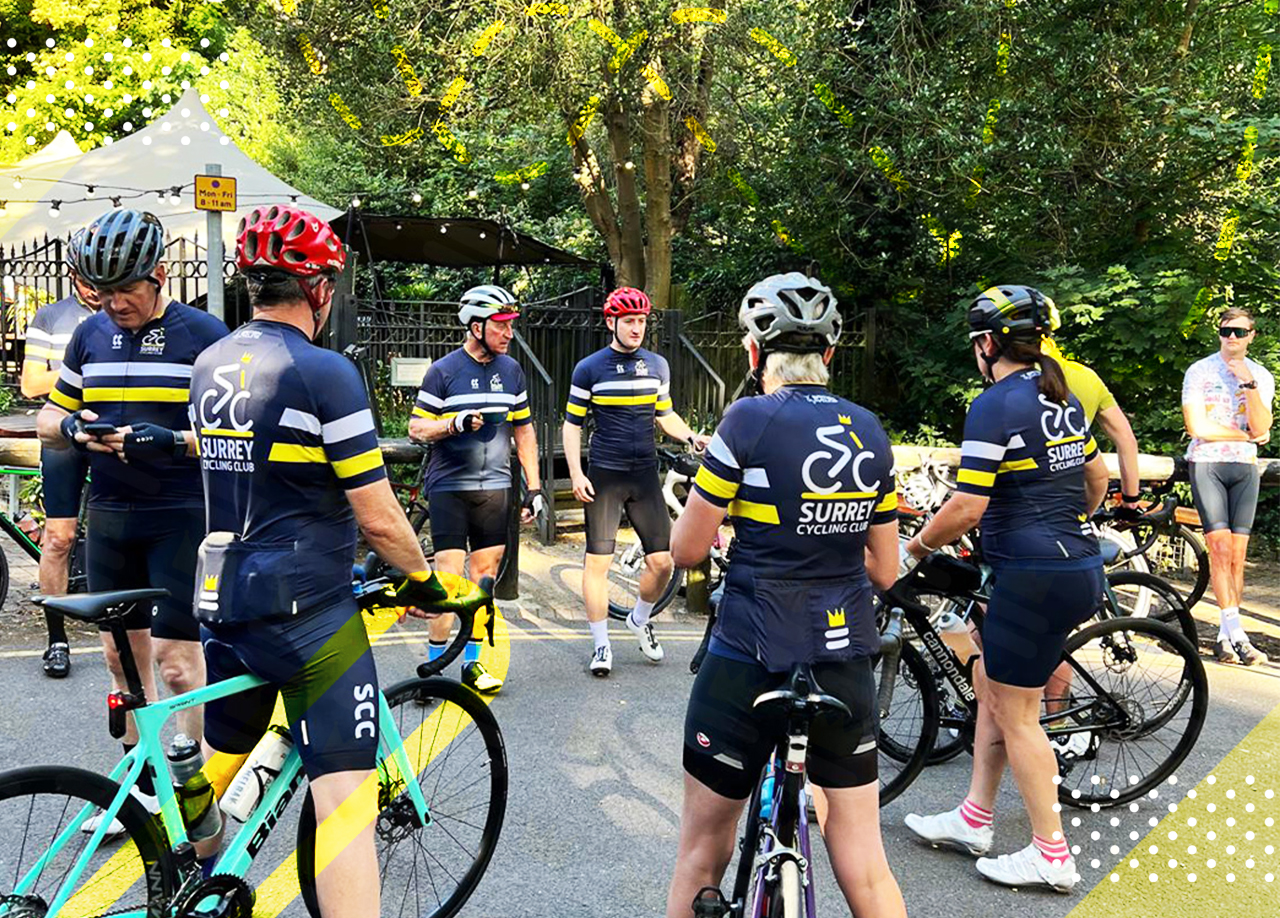 A group of cyclists in matching jerseys and helmets stand together with their bikes in an outdoor setting with greenery, preparing for a ride.
