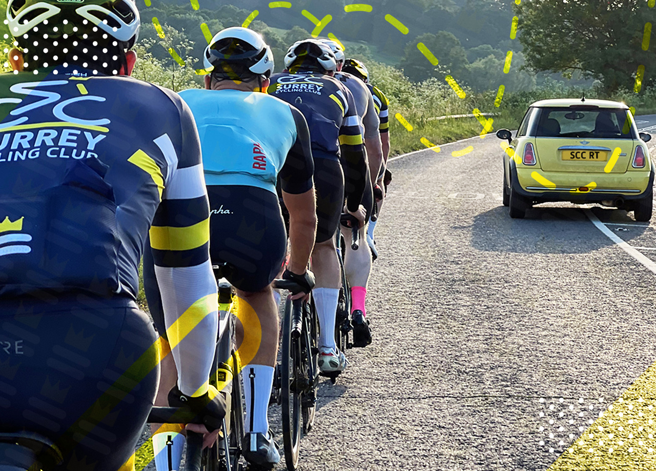 A group of cyclists from the Surrey Cycling Club ride in a single-file line on a sunny road, followed by a yellow car bearing the plate "SCC RT".